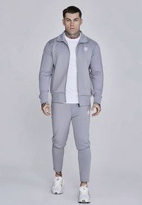 Tuta atletica grigia con giacca con zip e pantaloni affusolati. Texture liscia, logo sul petto, abbinata a scarpe da ginnastica bianche.