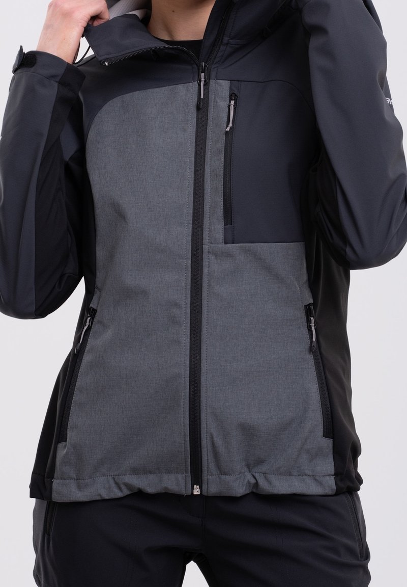 Personne portant une veste d'extérieur noire et grise avec fermeture éclair, avec des poches zippées sur la poitrine et les côtés, tenant la capuche d'une main.