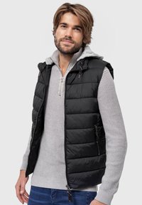 Sort kviltet vest med hætte, med grå detaljer, frontlynlås og sidelommer, båret over en lysegrå sweater i en afslappet setting.