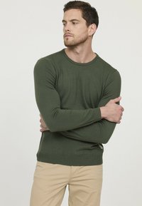 Pull en tricot vert à manches longues et col rond. Présente un design ajusté avec des poignets et un ourlet côtelés. Porté avec un pantalon beige.