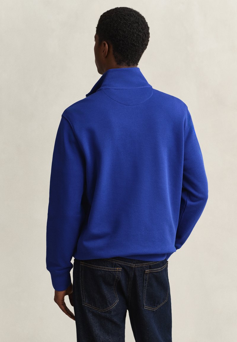 Man wearing a bright blue long-sleeve pullover with a high collar, paired with dark blue jeans, standing facing away against a plain background.