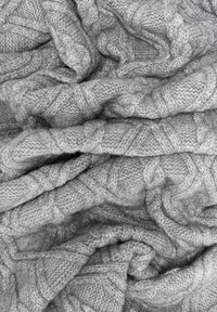 Szara, dziana kołdra o teksturowanym wzorze w warkocze, z miękkiej, grubej włóczki, częściowo złożona i przewieszona.