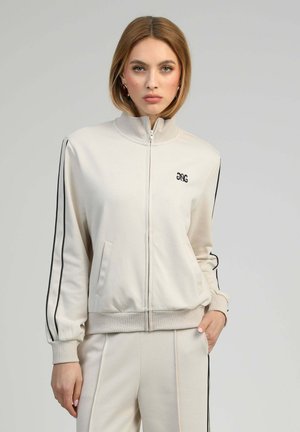 Femme portant un survêtement beige à fermeture éclair avec des rayures noires sur les manches et le pantalon, debout devant un fond clair uni.