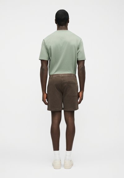 Homme debout de dos portant un t-shirt vert sauge, un short marron avec une poche arrière, des chaussettes blanches et des baskets blanches sur un fond uni.