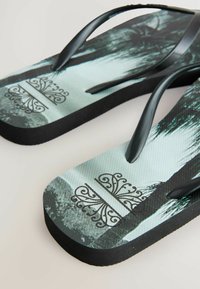 Sorte flip-flops med en struktureret sål, der har et palmetrædesign i grøn og grå, pyntet med udsøgte sorte grafikker nær midten.