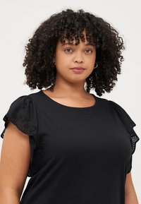 A woman with curly hair wears a black top with decorative sleeves, looking directly at the camera with a neutral expression.