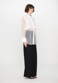 Transparent white blouse with a button-up front and a collar, featuring long sheer sleeves and contrasting white cuffs. Paired with black wide-leg trousers.