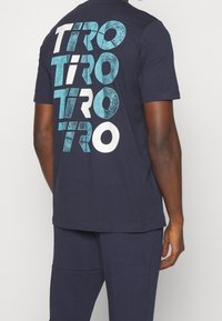 Námorně modré bavlněné tričko s velkým písmenem "TIRO" v tyrkysové a bílé barvě, s hladkou texturou a klasickým střihem, z pohledu zezadu.