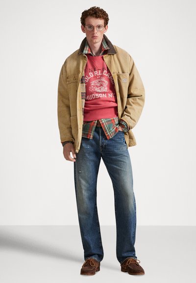Μπεζ μπουφάν καμβά πάνω από κόκκινο φούτερ με γραφικό σχέδιο, στρωμένο με πουκάμισο φανέλα καρό, μπλε τζιν και καφέ μοκασίνια.