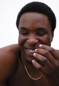 Nahaufnahme einer Person mit kurzen, lockigen schwarzen Haaren und einem hellbraunen Hautton, die eine goldene Kette trägt und mit sichtbaren Zähnen lächelt.