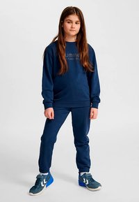 Mörkblå sweatshirt med ribbade muddar och nederkant, med en texturerad logotyp. Matchande mörkblå sweatpants med elastisk midjeband. Grå och blå sneakers.