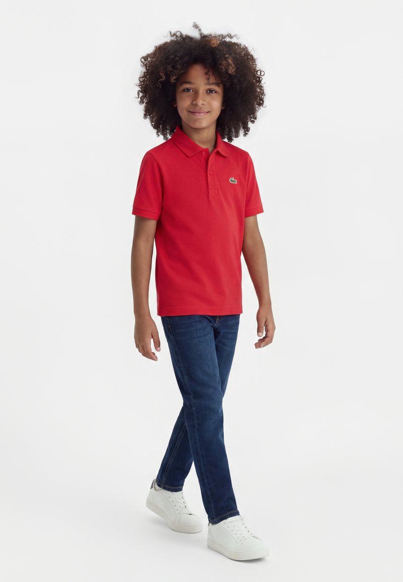 Enfant aux cheveux bouclés portant un polo rouge, un jean bleu et des baskets blanches, marchant avec un léger sourire sur fond blanc.