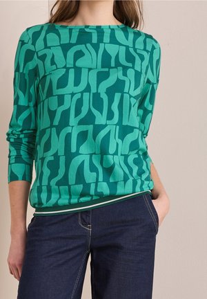 Femme portant un haut à manches longues vert à motifs et un jean bleu foncé, une main dans la poche, sur un fond beige.