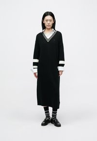 Črna dolga obleka z V-izrezom, belimi zvončastimi manšetami in izvezenim logotipom. Kombinirana s črnimi čevlji in črtastimi nogavicami.