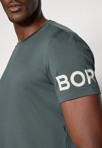 Dark green athletic t-shirt made of smooth fabric, featuring rounded neckline and a prominent white dotted print "BOR" on the sleeve.