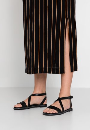 Person trägt schwarze flache Sandalen mit Knöchelriemen und einen schwarzen Rock mit vertikalen beigen Streifen und einem seitlichen Schlitz.