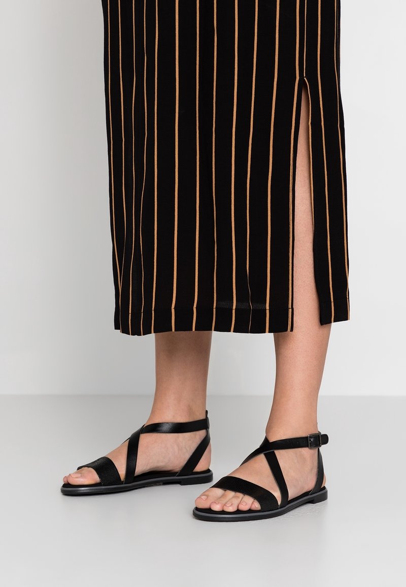 Personne portant des sandales plates noires avec des brides à la cheville et une jupe noire à rayures verticales beige avec une fente sur le côté.