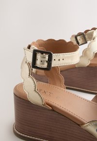 Sandale à talon compensé avec brides en cuir beige métallisé festonnées, boucle carrée noire et semelle intérieure rembourrée beige sur une semelle plateforme en bois.