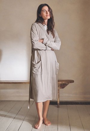 Frau steht barfuß auf einem Holzboden, trägt einen langen, gestreiften Bademantel mit Taschen, die Arme verschränkt, vor einer schlichten beigen Wand und einer Holzbank.