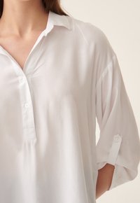 Blusa bianca con scollo a colletto, pattina con bottoni, maniche a tre quarti e polsini arrotolati. Tessuto morbido e liscio.