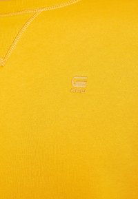 Sudadera amarilla con textura acanalada, que presenta un pequeño logotipo bordado en el lado izquierdo. Diseño simple con cuello redondo.