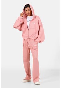 Sudadera rosa con cremallera, con bolsillos y pantalones rosa a juego, fabricados con un tejido suave. La sudadera cuenta con un cordón y un logo en la parte delantera.