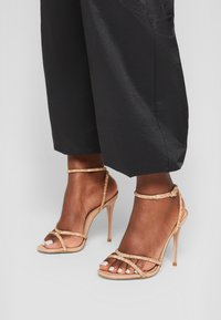 Hüfthochschnittige Nude-Stiletto-Sandalen mit einem Riemen-Design, das mit einem goldenen Glitzerakzent und einem Knöchel-Riemchen versehen ist. Veloursleder-Textur mit einer spitzen Zehenpartie.