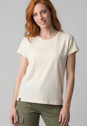 Rothaarige Frau, die ein schlichtes, off-white T-Shirt mit kurzen Ärmeln und olivgrüne Cargohosen trägt, leicht lächelnd vor einem grauen Hintergrund.