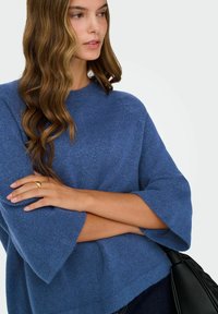 Femme aux cheveux bruns ondulés portant un pull en maille bleu à larges manches, les bras croisés, tenant un sac en cuir noir.