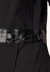 Vêtement en tissu noir avec une large ceinture en cuir noir présentant une grande boucle métallique de style épingle de sûreté à la taille.