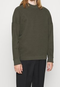 Sudadera de manga larga color verde oliva con cuello redondo, hombros caídos y puños acanalados. Tela suave con textura lisa.