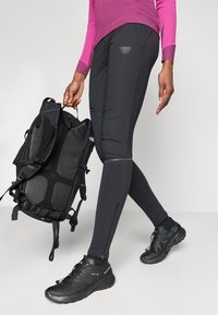 Zwarte sportleggings met een aansluitend ontwerp, zijritsen en een subtiele logodetail, gecombineerd met een zwarte rugzak en schoenen.