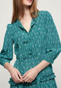 Blouse verte à motifs avec un design géométrique, un col et des détails de boutons. Manches longues avec des poignets élastiques et un ourlet volé.