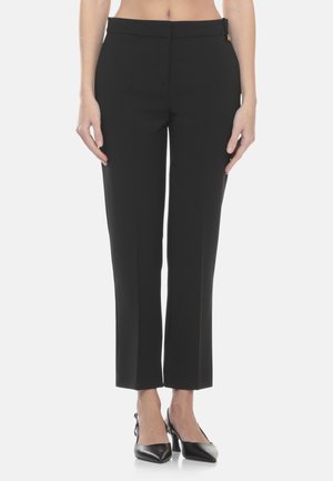 Donna che indossa pantaloni neri dritti alla caviglia abbinati a scarpe nere a punta con tacco e cinturino sul tallone, in piedi su sfondo bianco.