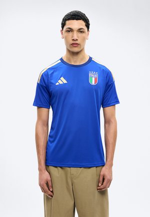 Giovane uomo che indossa una maglia da calcio dell'Italia blu con strisce e logo dorati, in piedi con espressione neutra su uno sfondo semplice.