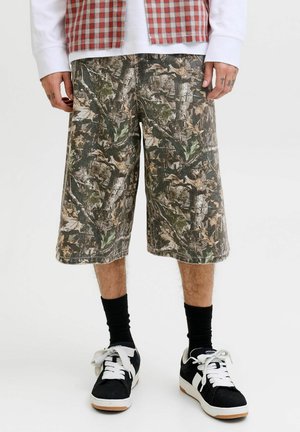 Mann iført knelang kamuflasjeshorts, svarte sokker og svarte og hvite joggesko står mot en ensfarget bakgrunn.