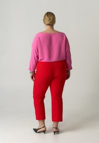 Blouse rose à col large et manches longues, associée à un pantalon rouge ajusté. Des escarpins noirs à bout pointu complètent la tenue.