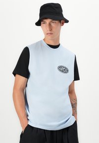 Ung man med svart bucket hat, svart t-shirt under en ljusblå ärmlös skjorta med en liten Rip Curl-logga, står med händerna i fickorna.