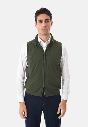 Uomo che indossa un gilet verde scuro con zip sopra una camicia bianca a maniche lunghe e jeans blu scuro, in piedi davanti a uno sfondo semplice.