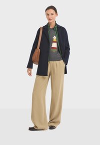 Abrigo de lana azul marino sobre una chaqueta acolchada verde, camiseta gris con un gráfico, pantalones de pierna ancha beige y bolso de hombro marrón. Zapatos oscuros.