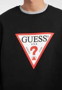 Melnas kokvilnas sporta džemperis ar lielu sarkanu un baltu trīsstūra logotipu, kurā ir teksts "GUESS U.S.A." un jautājuma zīmes dizains.