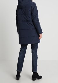 Abrigo acolchado azul marino con capucha, de diseño acolchado, de longitud hasta la rodilla y con mangas largas, combinado con jeans oscuros ajustados y botas hasta el tobillo.