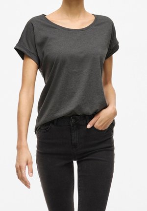 Personne portant un t-shirt gris foncé à manches courtes rentré dans un jean noir taille haute, avec une main dans une poche et l'autre détendue.