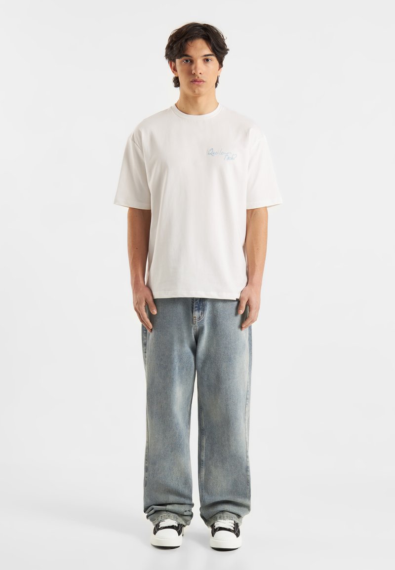 Witte katoenen t-shirt met een losse pasvorm, voorzien van lichtblauwe tekst. Gecombineerd met lichtblauwe baggy jeans en zwart-witte sneakers.