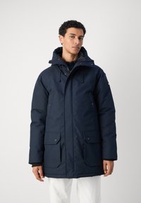 Navy blue parka jacket with a textured finish, adjustable hood, two front pockets, and a zip closure. Warm material designed for cold weather.