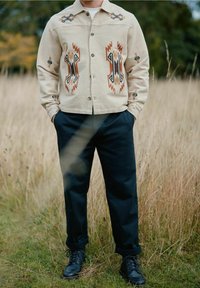 Beige jasje met ingewikkelde multicolore borduursels, met geometrische patronen. Gecombineerd met marineblauwe broek en zwarte veterschoenen.