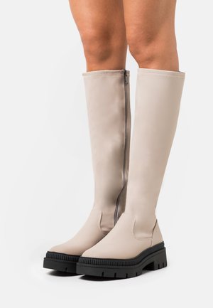 Bottes beige montant jusqu'au genou avec une finition lisse, une fermeture éclair latérale et des semelles en caoutchouc noir épaisses avec un motif de bande de roulement texturé.