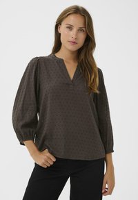 Bruine textuur blouse met een V-hals, pofmouwen en subtiele verticale patronen. Draagt met zwarte broek, handen in de zakken.