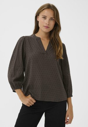 Bruine textuur blouse met een V-hals, pofmouwen en subtiele verticale patronen. Draagt met zwarte broek, handen in de zakken.