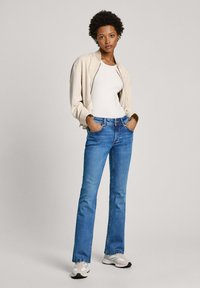 Una chaqueta bomber beige claro sobre una blusa blanca ajustada, combinada con vaqueros acampanados azules. El modelo está de pie con las manos en los bolsillos, usando zapatillas ligeras.
